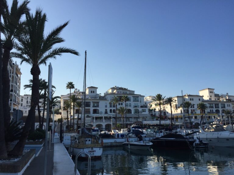 Boats moored at Puerto de la Duquesa in Manilva, with white Andalusian apartment buildings and palm trees behind the marina
